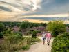 Zwei Wanderer auf einem sandigen Weg durch Heidelandschaft mit blühender Heide und Büschen unter bewölktem Himmel.