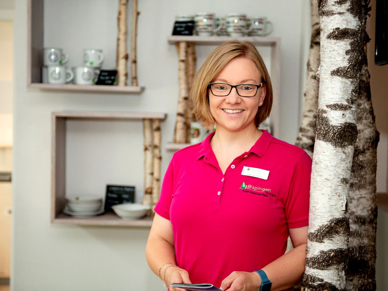Franziska Kühne von der Tourist Information Bispingen lächelnd, trägt pinkes Poloshirt mit Logo, steht neben Birkenstamm in hellem Raum
