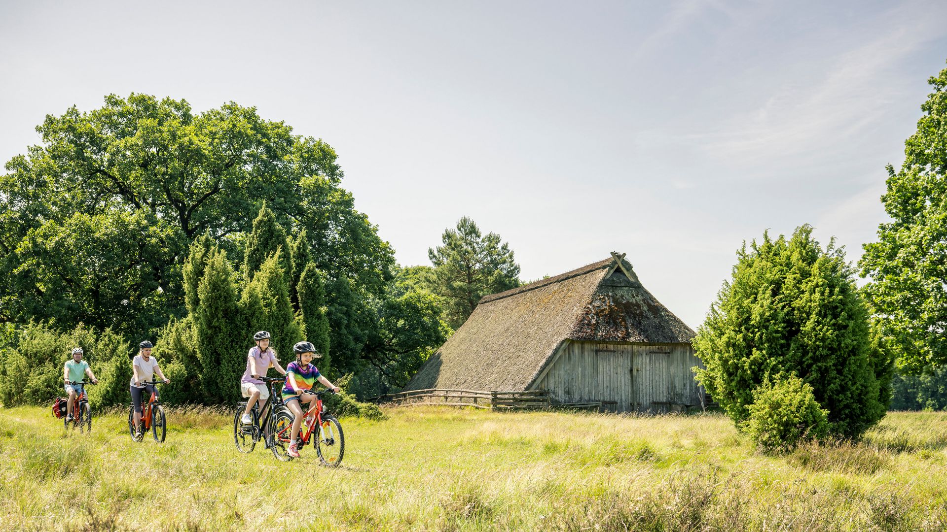 Familie fährt mit Fahrrädern auf einem Weg im Naturschutzgebiet bei Bispingen, vor einer traditionellen Reetdachscheune in der Lüneburger Heide