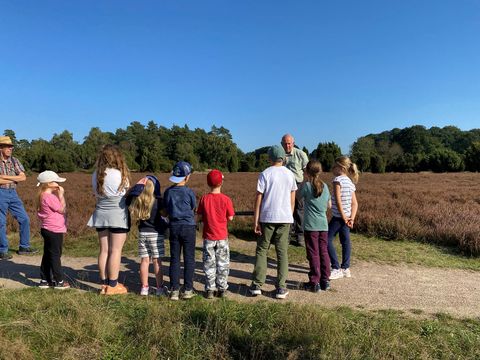 Gruppe von Kindern steht auf einem Weg vor einer Heidelandschaft, ein Erwachsener spricht zu ihnen, umgeben von Bäumen unter klarem Himmel