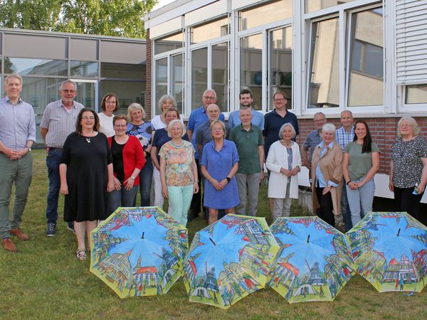 Personengruppe vor einem Haus mit aufgespannten Regenschirmen auf dem Boden.