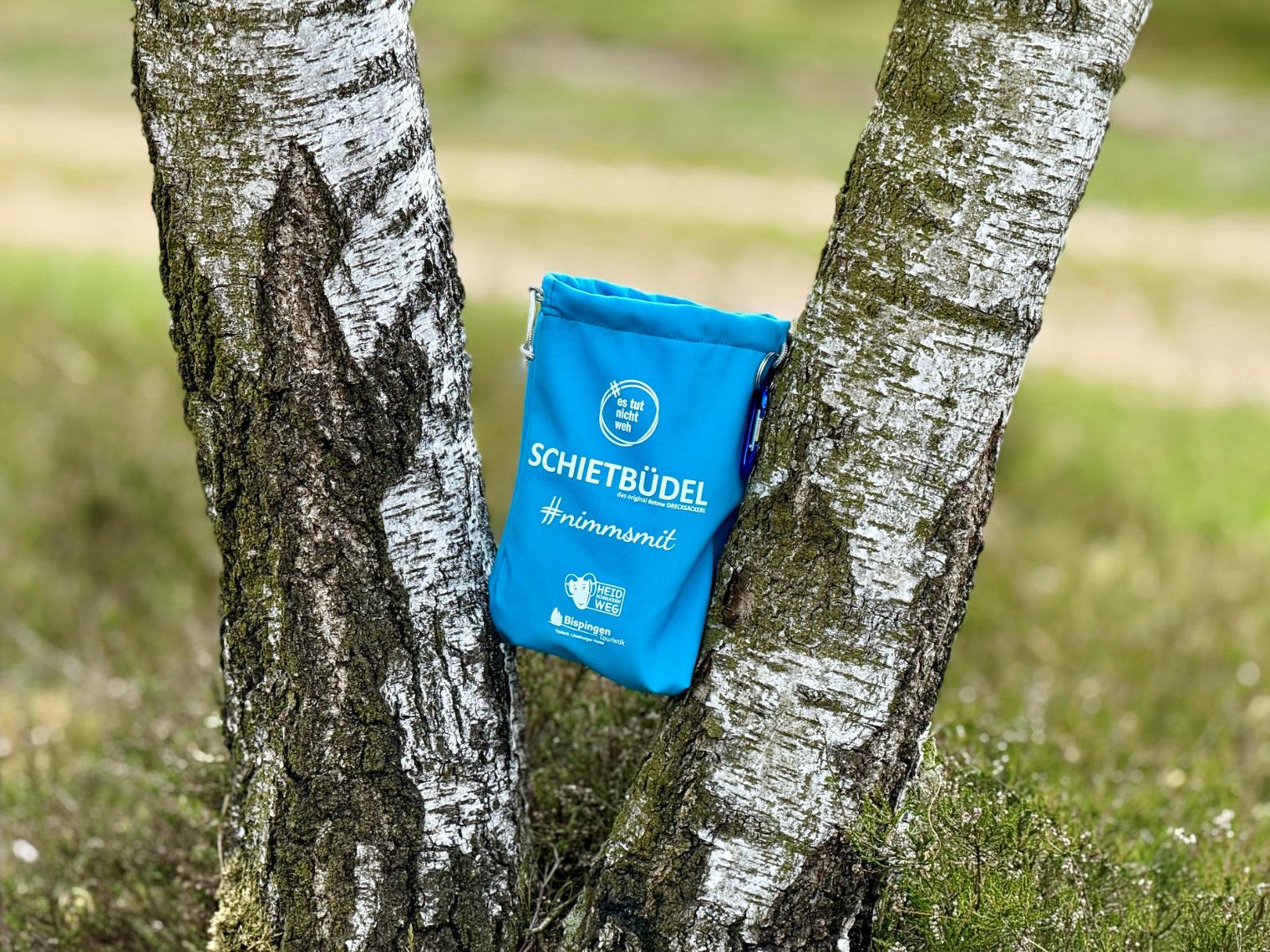 Blauer "Schietbüdel" Müllbeutel hängt zwischen zwei Birkenstämmen in der Heide, mit Aufschrift 'Schietbüdel #nimmmit' und Logos von Bispingen Touristik und Heidweg