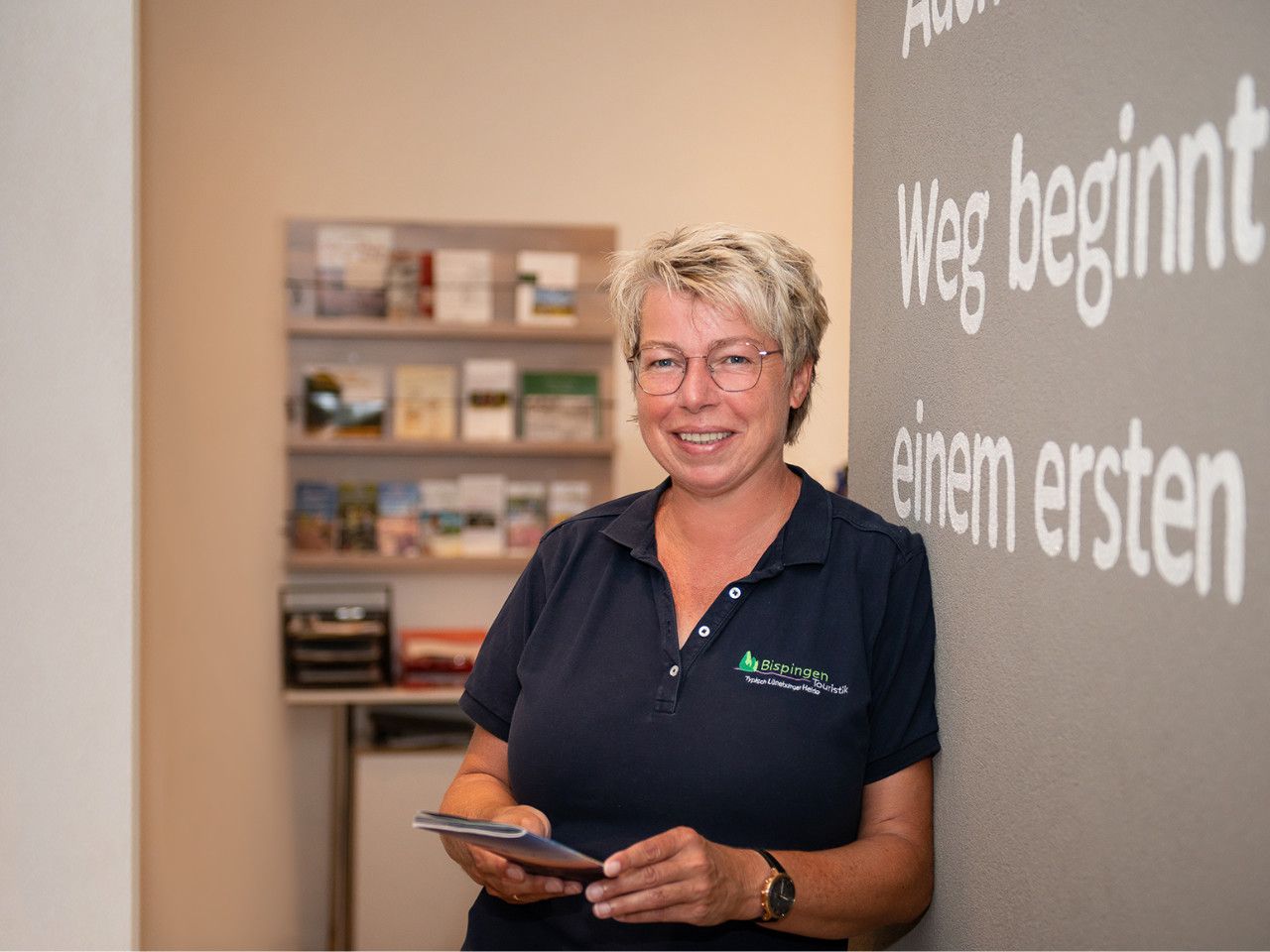 Mitarbeiterin Anja Meyer der Tourist Information Bispingen steht lächelnd mit Tablet vor einer Wand mit Text