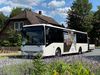 Weißer Heide Shuttle Bus fährt auf einer Straße vor einem Fachwerkhaus, umgeben von grünen Bäumen und bunten Blumen unter blauem Himmel mit weißen Wolken