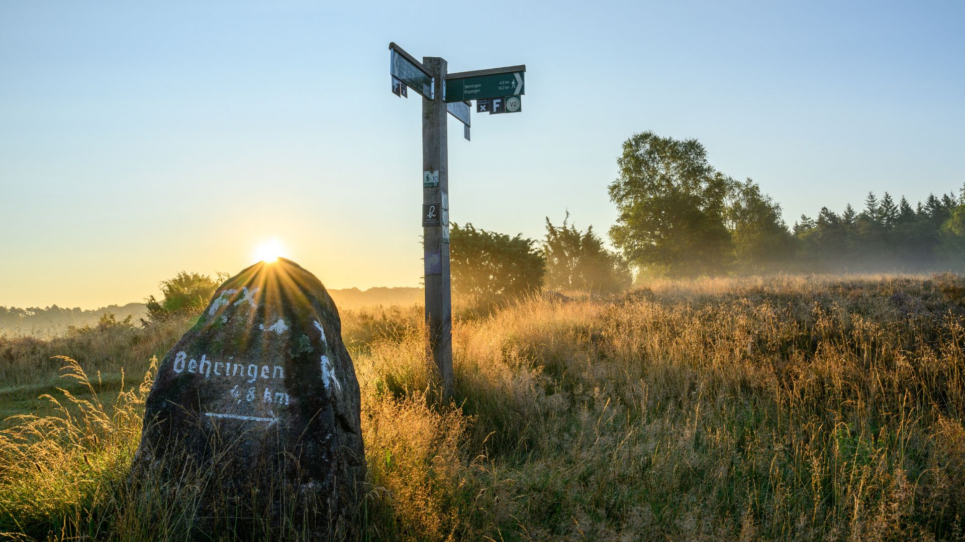 Sonnenaufgang über einer Heidelandschaft am Heidschnuckenweg bei Bispingen mit Wegweiser und Stein mit der Aufschrift 'Behringen 8 km'