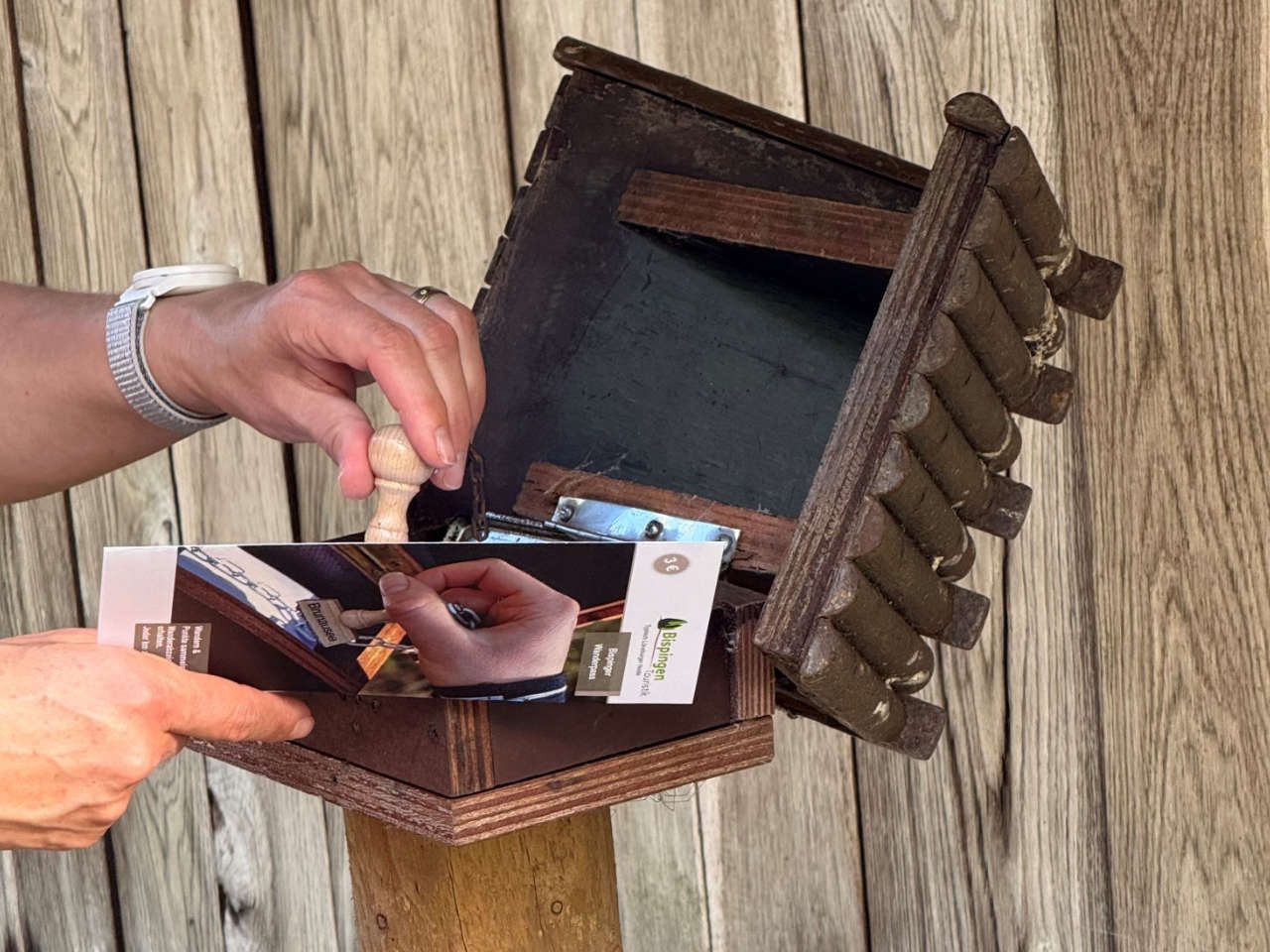 Hände stempeln einen Wanderpass vor einem hölzernen, kleinen Häuschen mit Stempelkissen
