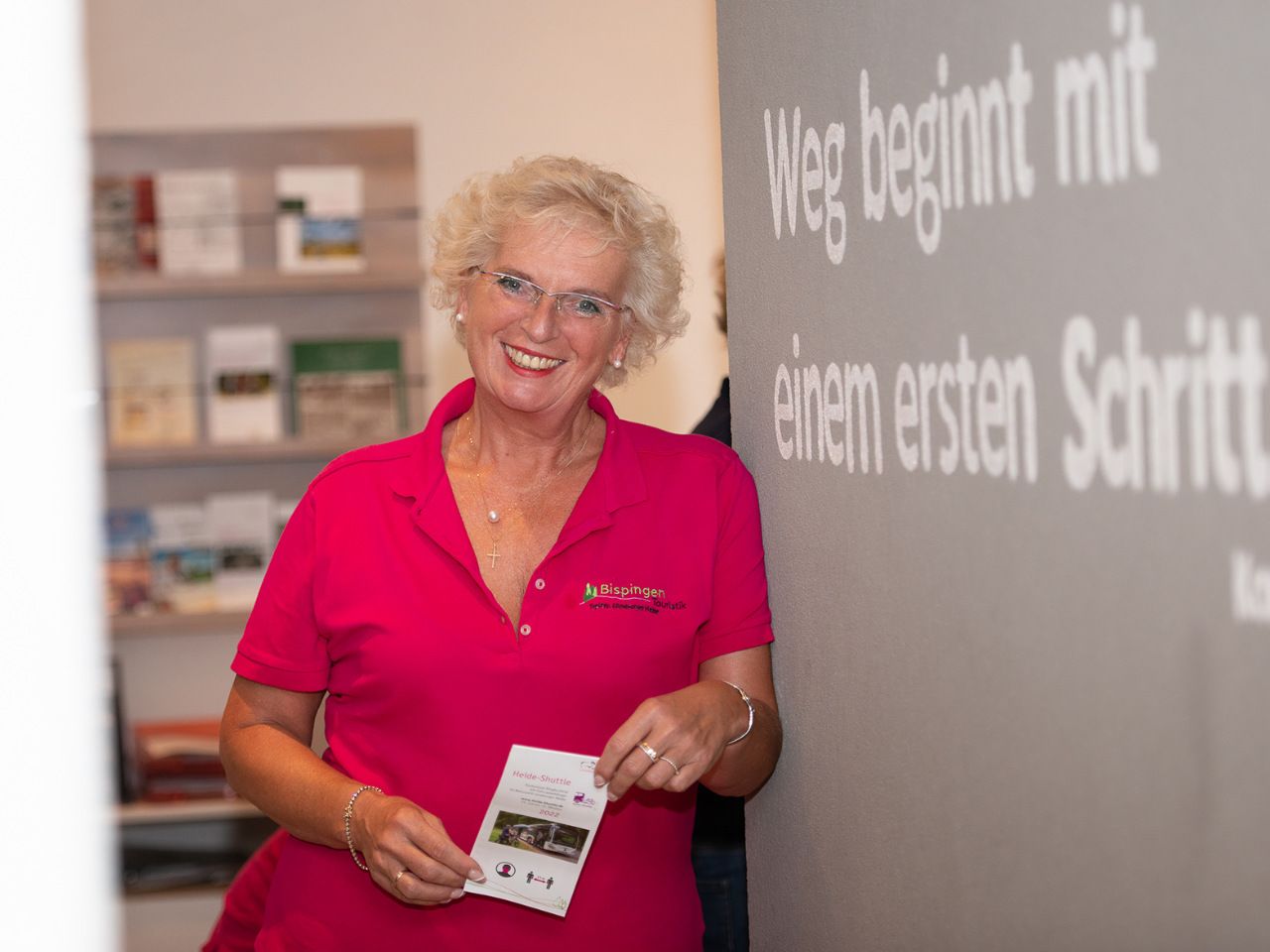 Mitarbeiterin Silke Rudat der Tourist Information Bispingen lächelnd im pinken Poloshirt, hält Flyer, steht vor Wand mit Schriftzug 'Weg beginnt mit einem ersten Schritt'