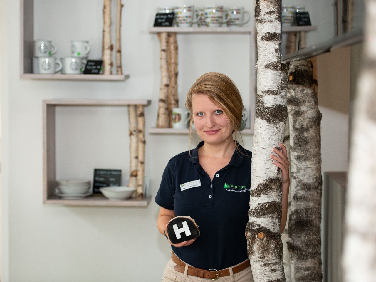 Mitarbeiterin Sarah Schönfelder der Tourist Information Bispingen in dunkelblauem Poloshirt mit Logo, lächelnd, neben einer Birkenholzsäule