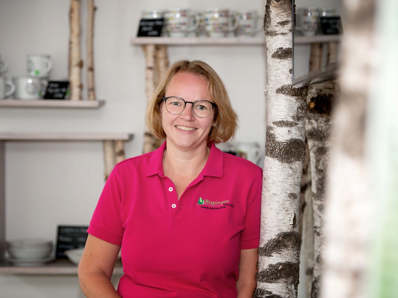 Mitarbeiterin Catrin Sorge der Tourist Information Bispingen lächelt, trägt ein pinkes Poloshirt mit Logo und steht vor einem Regal mit Geschirr in einem hellen Raum