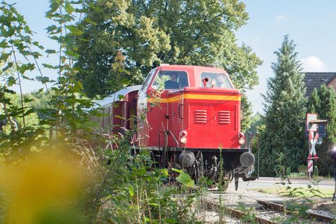 Historische rote Diesellokomotive auf Gleisen in ländlicher Umgebung mit Bäumen und Häusern im Hintergrund