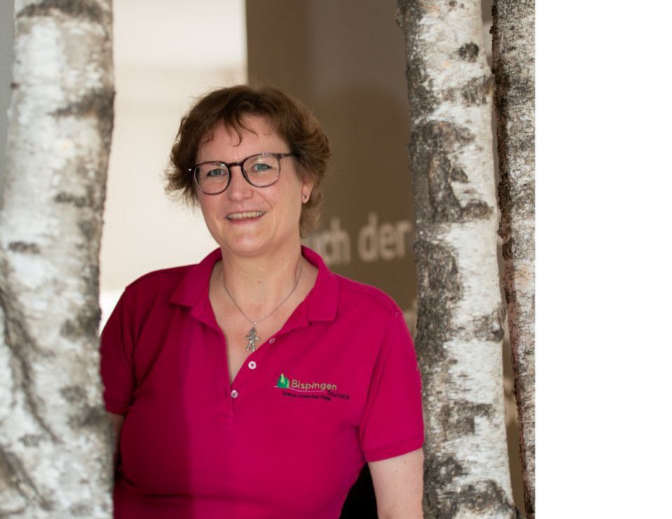 Christine Wortmann von der Tourist Information Bispingen steht zwischen zwei Birkenstämmen, trägt ein pinkes Poloshirt mit Bispingen-Logo und lächelt freundlich